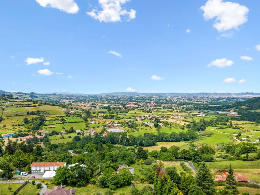 Vista panorámica de viviendas y prados, con la ciudad al fondo.