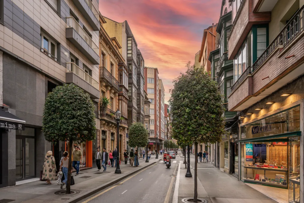 Calle arbolada con edificios, comercios y peatones bajo un cielo rosado al atardecer.