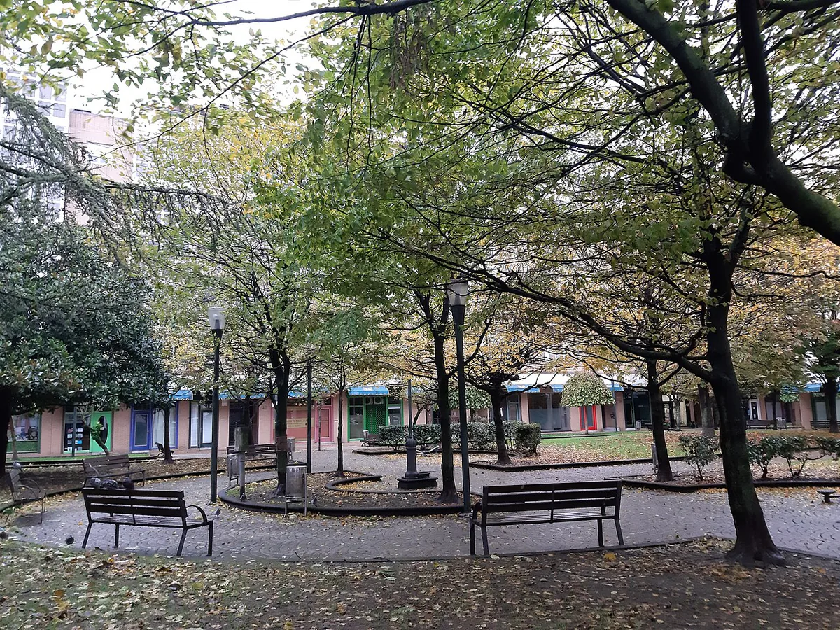 Parque con árboles, bancos y farolas, con hojas caídas sobre los caminos y locales al fondo.