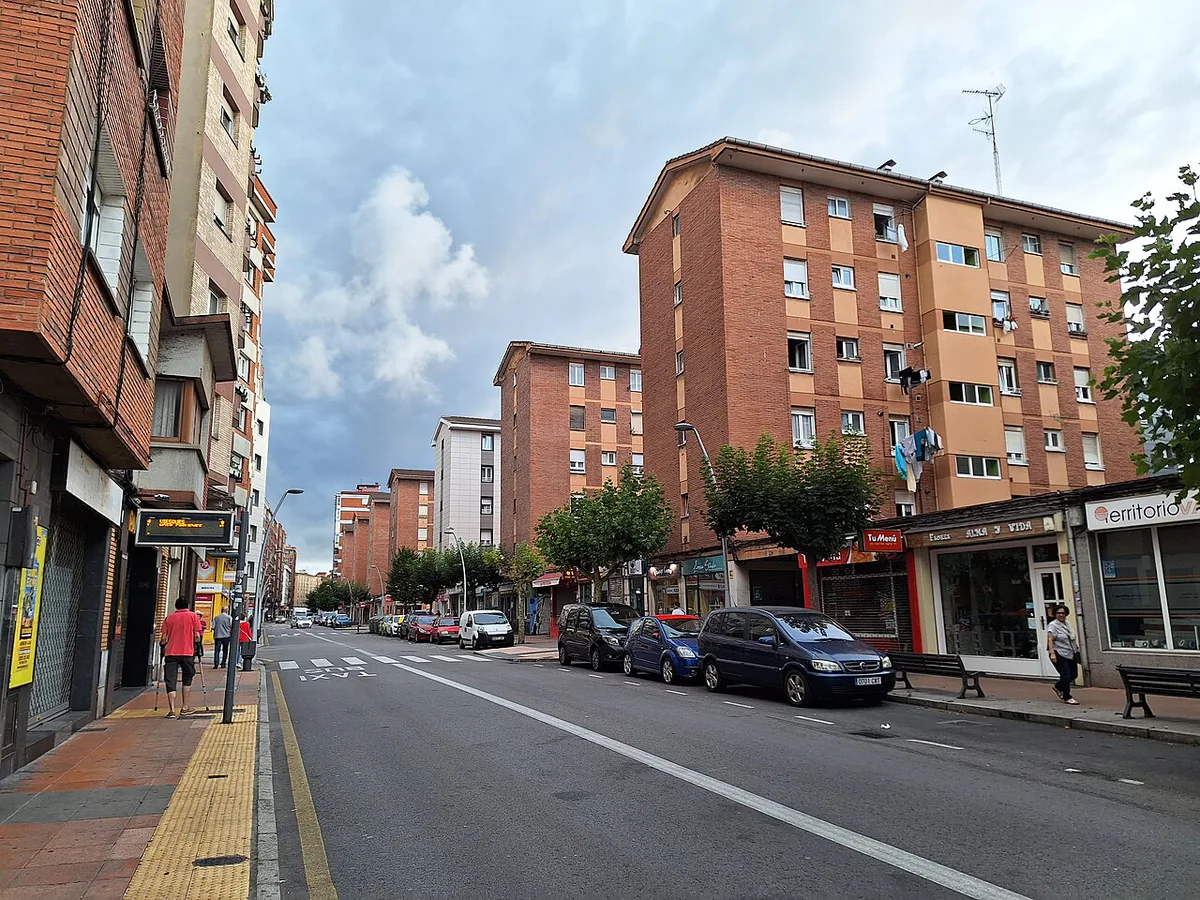 Calle con edificios de viviendas, comercios, árboles, bancos y un carril señalizado para autobús y taxi.
