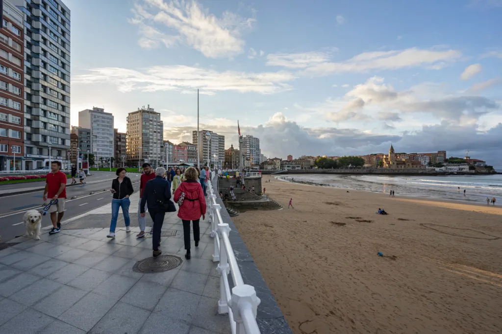 Paseo junto a la playa, con peatones, una barandilla blanca y edificios al fondo.