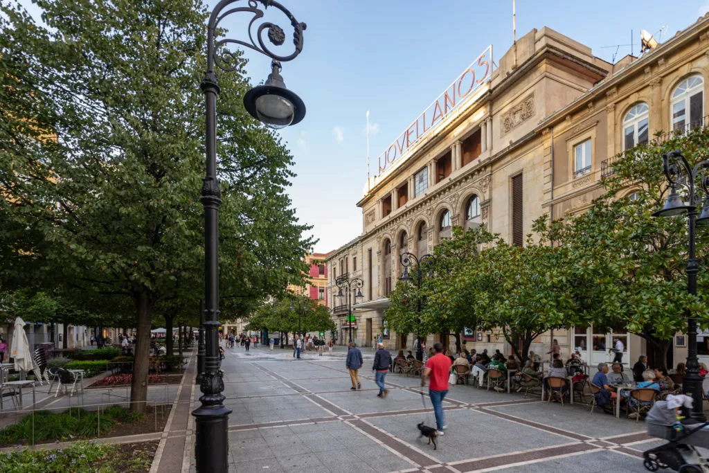 Fachada del Teatro Jovellanos junto al paseo arbolado de Begoña, con farolas y peatones.